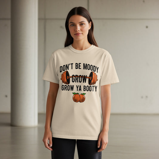 Woman wearing cream t-shirt with "Don't be moody, grow ya booty" text and barbell graphic.
