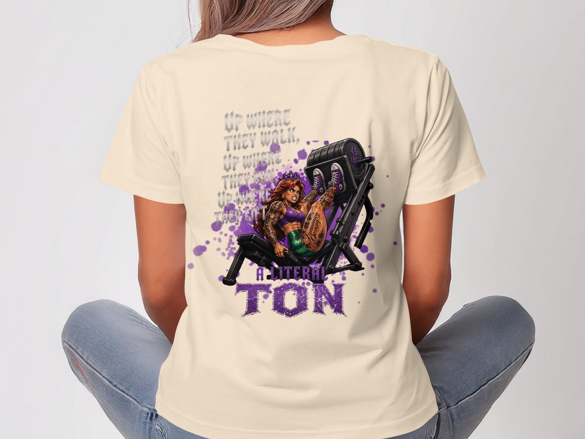 Woman wearing a cream t-shirt with a purple graphic of a woman on a weight machine.