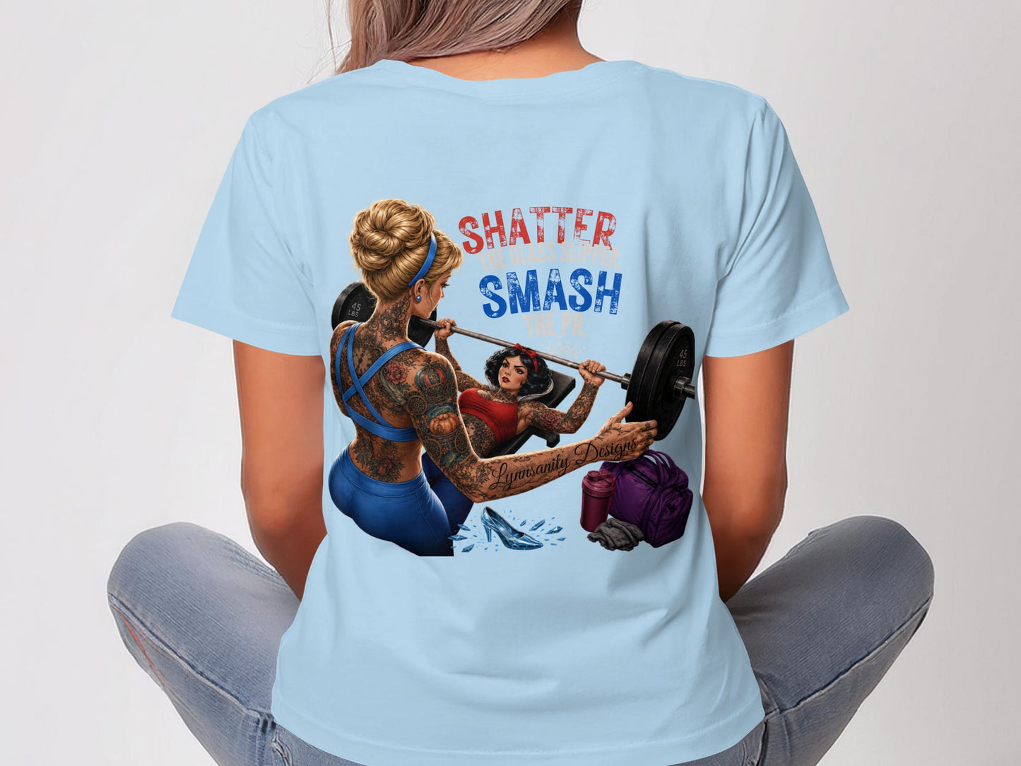 Woman in light blue t-shirt with "SHATTER SMASH" graphic and barbell workout scene.