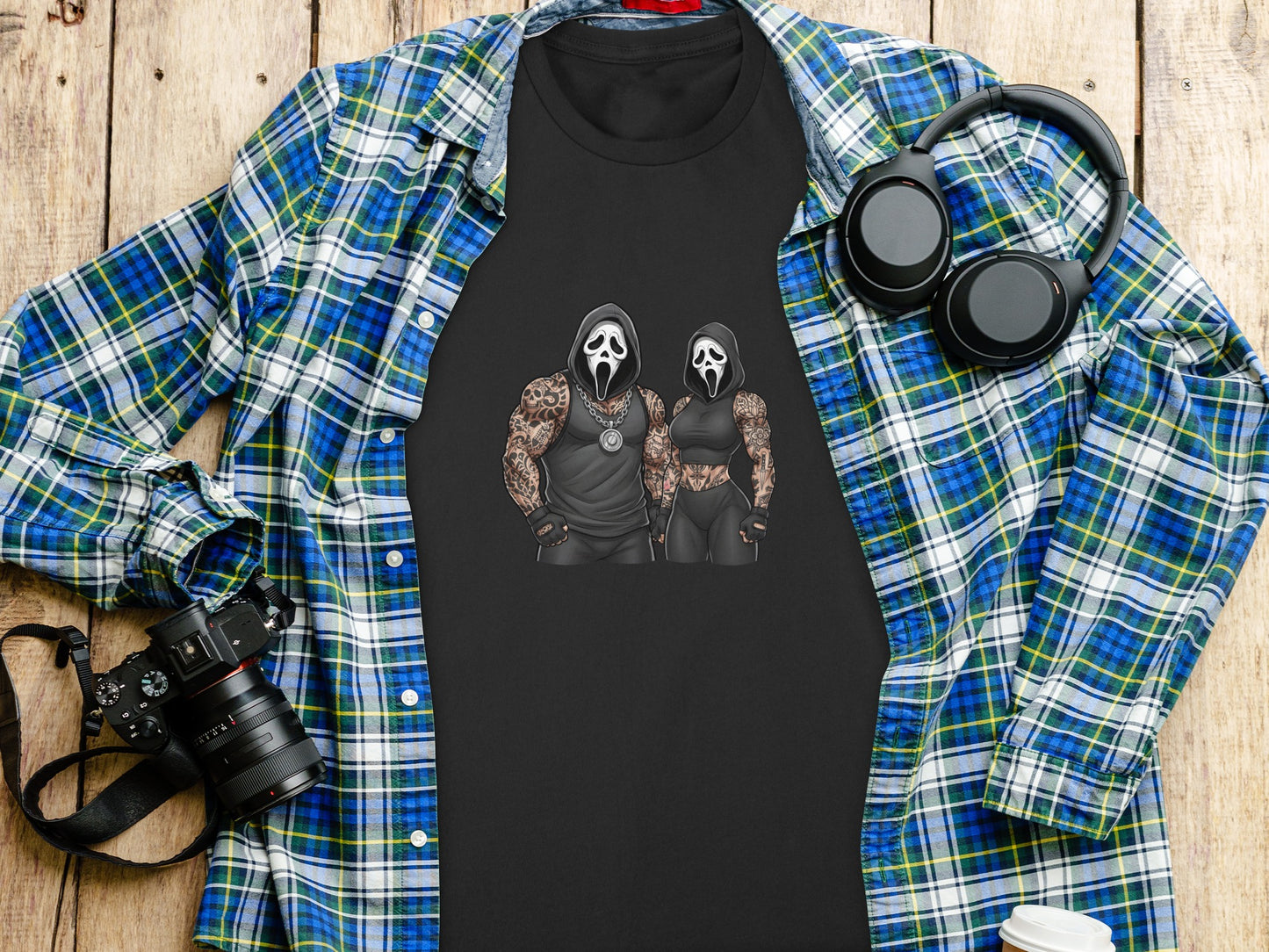 Black t-shirt with Ghostface couple graphic, spread on a wooden surface with camera and headphones.