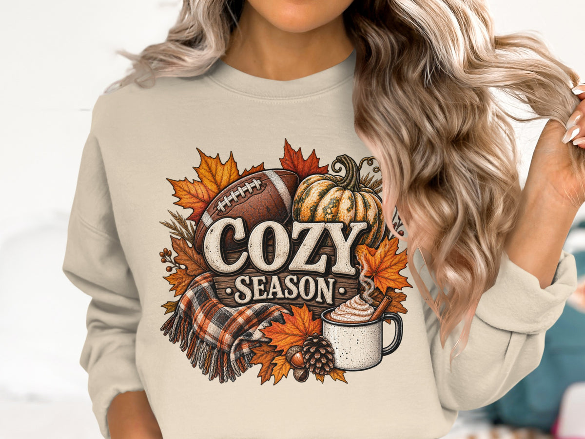 Crewneck sweatshirt with "Cozy Season" graphic featuring football, pumpkin, plaid scarf, and autumn leaves.